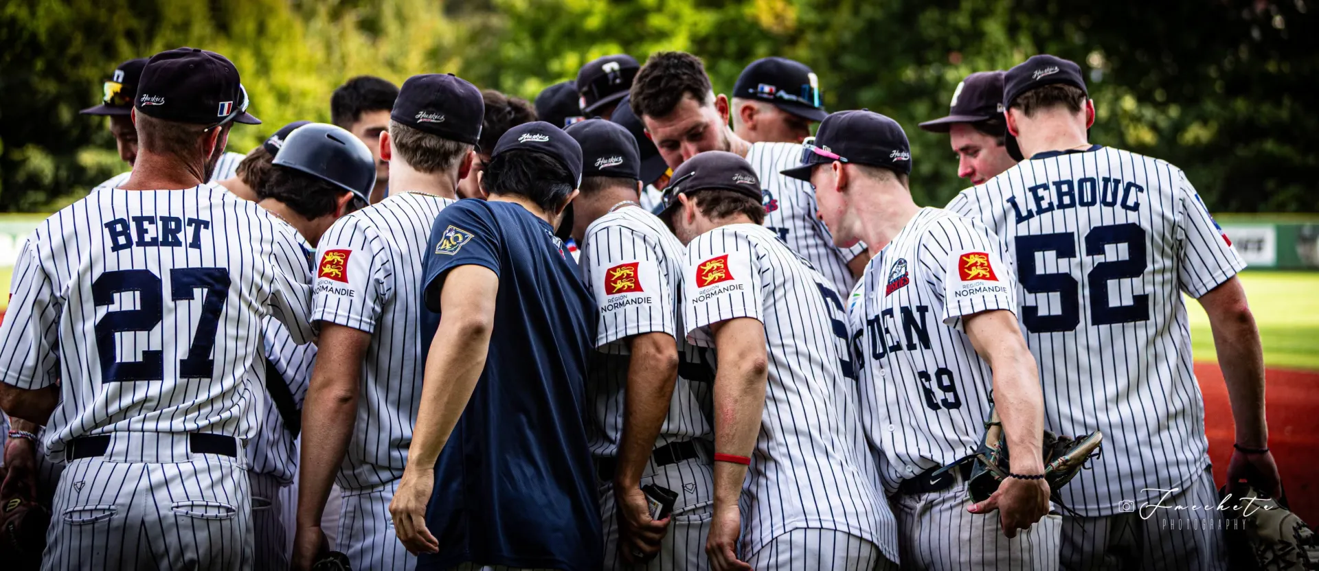 Les joueurs des Huskies de Rouen, maillots à fines rayures et casquettes marine, regroupés en cercle sur le terrain lors d'un match de Division 1 2026.