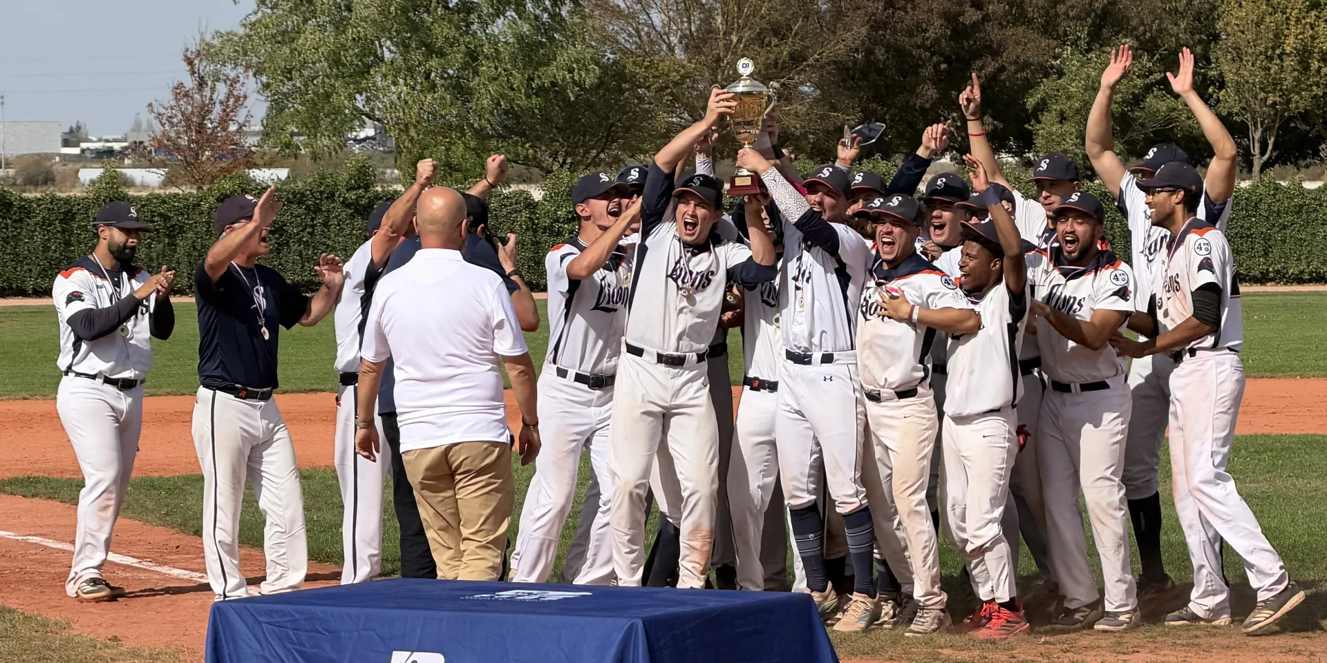 Les joueurs des Lions de Savigny, maillots blancs et casquettes marine au S, brandissent le trophée de champion de France en criant de joie, groupés autour du marbre du C'Chartres Baseball Softball, devant une table aux couleurs de la FFBS.